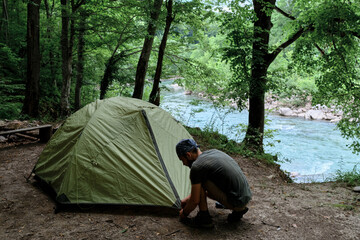 Put tourist tent in campsite and prepare for rest. Young handsome Caucasian male traveler with bandana on his head and beard sets up tent for rest in clearing in forest.