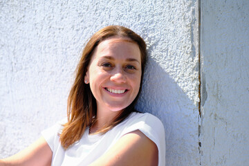young adult woman smiling and looking at camera. freckled caucasian woman sitting next to an old house wall and enjoying summer