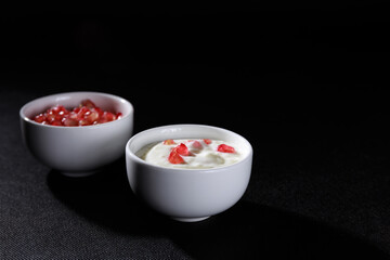 White bowl with lactose-free yogurt and other bowl with pomegranate grains, on black background