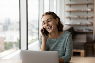 Joyful young woman involved in pleasant mobile phone call talk, distracted from computer work in office. Smiling millennial business lady discussing working issues distantly, multitasking concept.