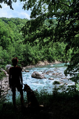 Obraz premium Viewing platform on mountain and female tourist. Traveler girl and German Shepherd dog stand together on cliff and look at stormy beautiful mountain river flowing at bottom of stone gorge.