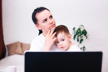 a young black-haired mother with a small child works at home in a bright room with a laptop.