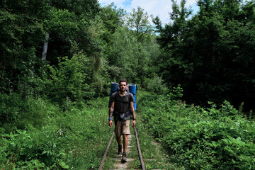 Traveler guy with big backpack walks along rails and sleepers forward. Young handsome Caucasian man with beard and dreadlocks is walking along railway among green summer forest.