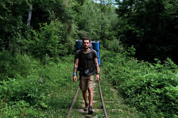 Obraz premium Traveler guy with big backpack walks along rails and sleepers forward. Young handsome Caucasian man with beard and dreadlocks is walking along railway among green summer forest.