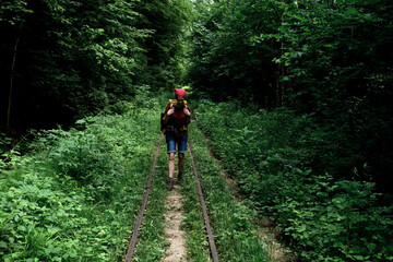 Fototapeta na wymiar Woman tourist explores new places and trails. Caucasian female traveler is walking along railway with large yellow backpack among green summer forest. Front view.