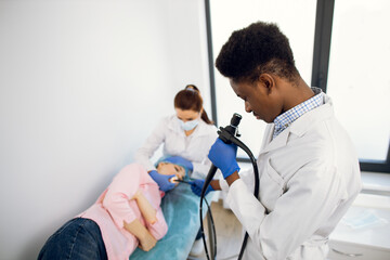 Close up of male Afro-American doctor with an endoscope and female colleague nurse examining young woman patient. Endoscopy, medical examination