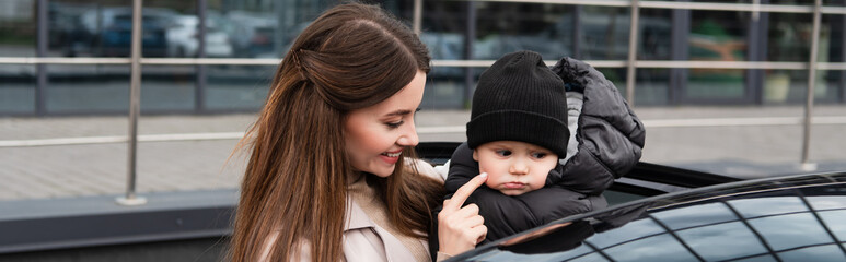 smiling woman touching cheek of baby boy near car on urban street, banner