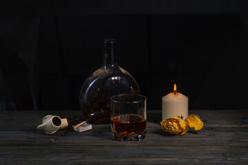 An old dirty drink bottle, a filled glass, a burning candle, a dried yellow rose and a smoking pipe...