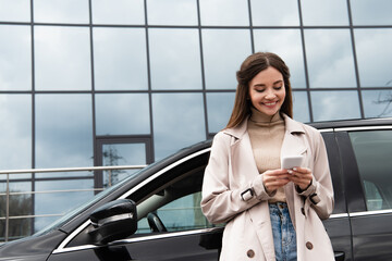 cheerful woman in trench coat using smartphone while standing near car