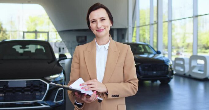 Beautiful caucasian female seller holding clipboard, smiling to the camera. Successful saleswoman standing in front of modern vehicle demonstrates shows car and looks in camera. Concept of car selling