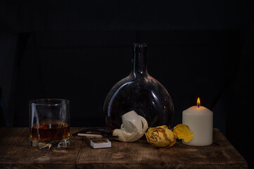 An old dirty drink bottle, a filled glass, a burning candle, a dried yellow rose and a smoking pipe on a wooden table on a dark background with a bokeh effect