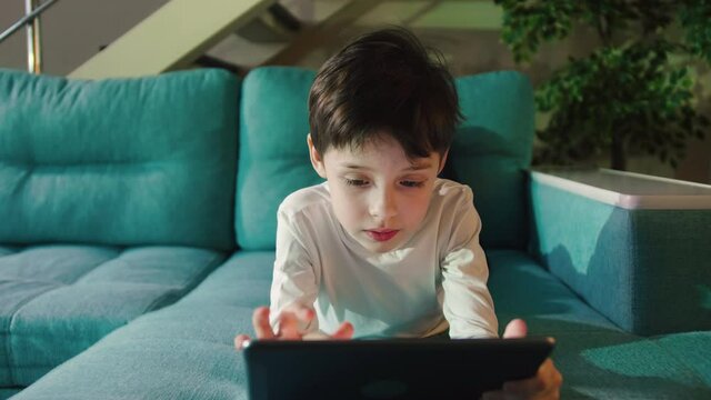 Close Up Young Boy Lying In Bed And Using Digital Tablet. Young Schoolboy Staring At Pad Computer Watching Video At Home. Concept of Information Overload, Virtual Smartphone Addiction