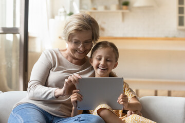 Happy loving middle aged granny resting on comfortable sofa with joyful adorable preschool kid girl, using digital computer tablet together, watching funny photo video content or playing online games.