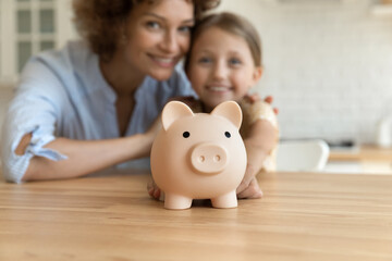 Close up focus on piggybank standing on table with blurred happy mother and little kid daughter on background. Friendly smiling joyful two generations family planning future investment or saving money