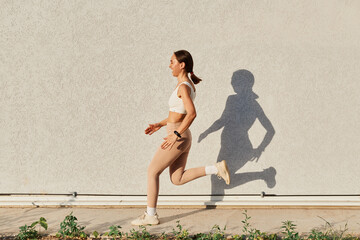 Side view full length portrait of laughing female wearing white top and beige leggins running outside, dark haired woman with ponytail doing with perfect body.