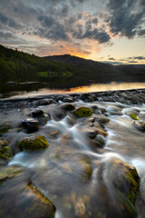 Flowing water from weir with beautiful scenic view of Grasmere lake in the Lake District, UK. Dramatic sunset sky in background with reflections.