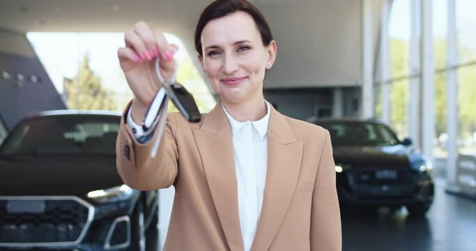 Portrait of handsome car dealer in formal suit holding keys from new car. Female dealer standing at auto salon, smiling and looking at camera.