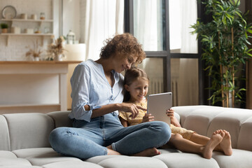 Relaxed carefree affectionate young mother and little preschool child daughter sitting on cozy couch, playing games on digital touchpad, spending leisure weekend time using educational applications.