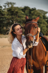 Cute happy young woman with horse in summer beach by sea. Rider female drives her horseback in nature on evening sunset light background. Concept of outdoor riding, sports and recreation. Copy space