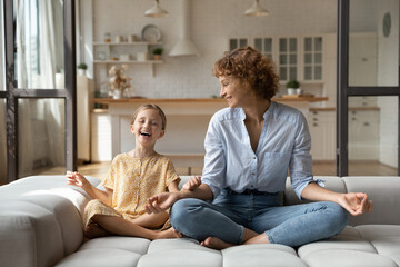 Happy adorable small kid girl sitting in lotus position with caring beautiful young mother, having fun learning meditating together on comfortable couch, enjoying carefree leisure weekend time.