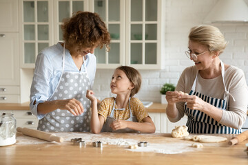 Obraz na płótnie Canvas Happy young 35s woman talking enjoying cooking homemade pastry with joyful cute little preschool kid daughter and caring beautiful older retired mother in modern kitchen, family culinary activity.