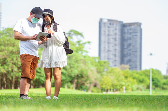 Asian couples with facial mask searching for tourist information through tablets of amazing places to visit in urban with building and park background. Technlogy and Travel concept. New normal theme
