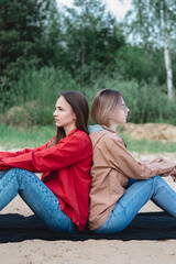 Two young caucasian young women friends sitting back to back on the sand at the beach