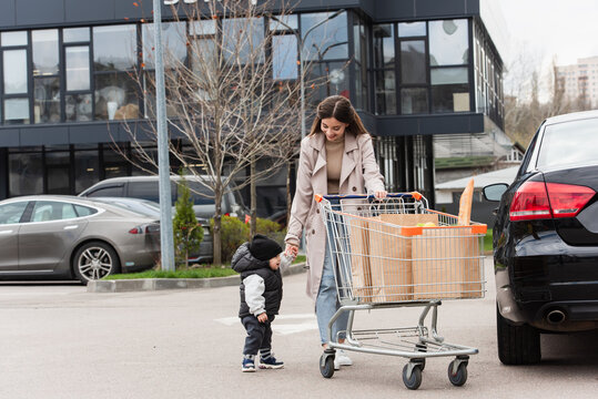 Mother With Toddler Son Walking On Car Parking Near Shopping Trolley