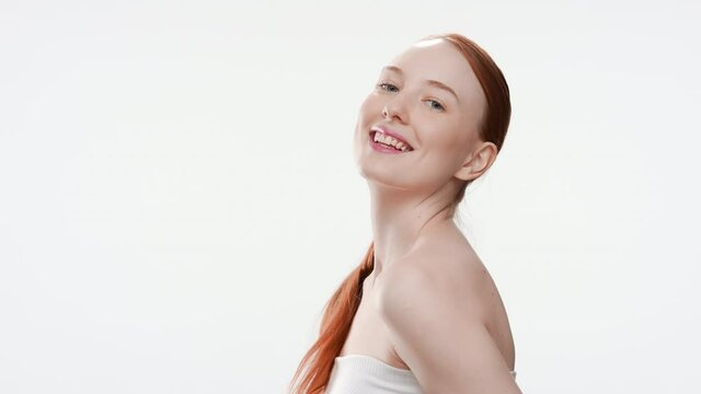 Young gorgeous fit Caucasian woman with long ginger hair in ponytail wrapped in a towel turns around smiling wide for camera on white background | Pure skin concept