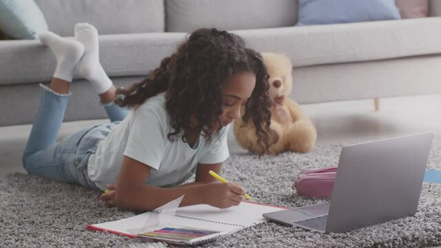 Cute african american girl drawing with felt-pens, lying on floor with laptop, watching online art lessons, slow motion