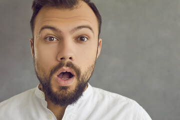 Closeup studio portrait of surprised bearded young man gazing open mouthed at camera. Close up shot of handsome guy looking at you with shocked face expression