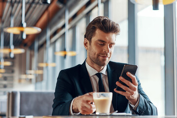 Handsome young man in full suit using smart phone and drinking coffee while sitting in the restaurant