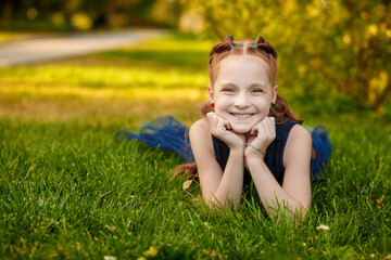 Naklejka na ściany i meble A beautiful little redhead girl in a blue dress lies on the grass in a city park on a warm summer day. Children's emotions of joy. Space for text. Funny photo.