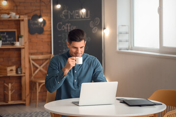 Handsome young man drinking coffee while working with laptop in cafe