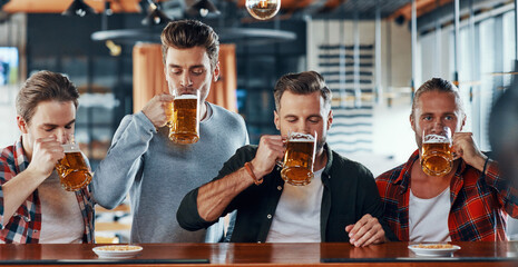 Group of handsome young men in casual clothing enjoying beer while sitting at the bar counter in pub
