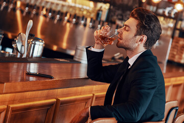 Handsome young man in full suit having alcohol drink while sitting at the bar counter in restaurant