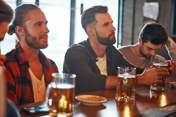 Group of young men in casual clothing enjoying beer while watching TV in the pub