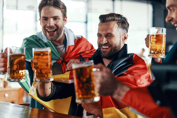 Happy young men covered in international flags toasting with beer while watching sport game in the pub