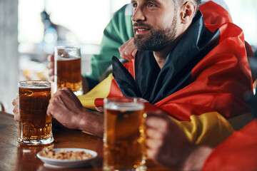 Young men covered in national flags enjoying beer while watching sport game in the pub