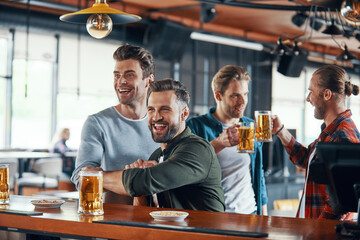 Cheering young men in casual clothing watching sport game and enjoying beer while sitting in the pub