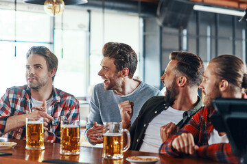 Cheering young men in casual clothing enjoying beer while sitting in the pub