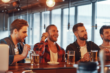 Group of happy young men in casual clothing enjoying beer while sitting at the bar counter in pub