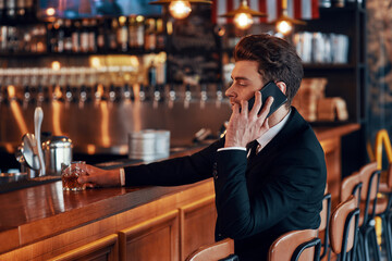 Handsome young man in full suit talking on smart phone while sitting at the bar counter in restaurant