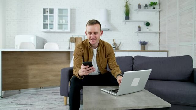Smiling freelancer with a phone on kitchen background. Young man student using a laptop and mobile phone studying online. Guy working from home enjoying distant job.