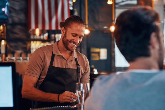 Young male bartender cleaning glasses and smiling while standing at the bar counter in pub