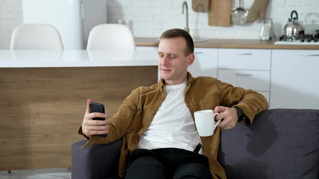 Handsome man using a phone at home. Guy rests on sofa with a cup of coffee and doing photos on a smart phone in the kitchen.
