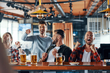 Cheering young men in casual clothing drinking beer and watching sport game while sitting in the pub
