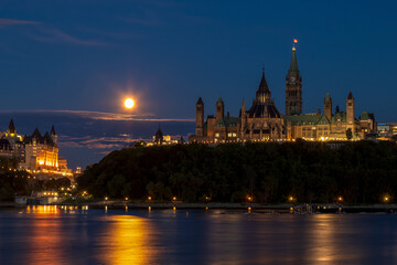 Naklejka premium Downtown Ottawa at night by the river. Full moon shining low in the blue sky. Parliament buildings and historic hotel in Ottawa, Ontario, Canada