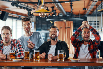 Cheering young men in casual clothing drinking beer and watching sport game while sitting in the pub