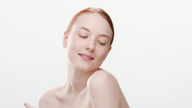 Close-up beauty portrait of beautiful fit Caucasian young red-haired woman with naked shoulders touches her face averting her eyes down on white background | Face care concept
